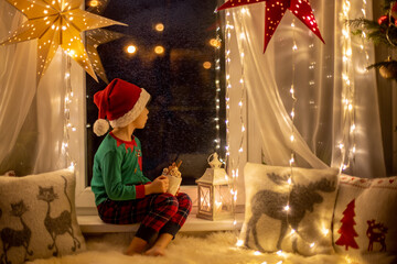 Toddler child, cute blond boy, sitting on the window, reading book, Christmas lights around