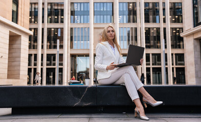 Upset young blonde entrepreneur woman using laptop with sad facial expression  sitting outdoors against large building. Pretty student girl preparing for exam. Overloaded businesswoman typing letter.