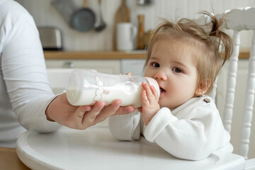Baby toddler sit in high chair in kitchen and drink milk or formula from bottle. Mom's hand helping to feed child