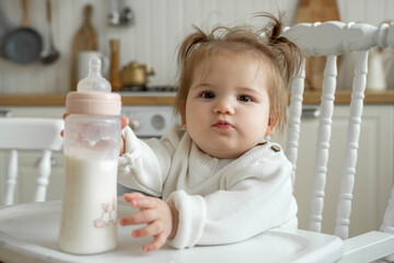 Baby toddler sit in high chair in kitchen and drink milk or formula from bottle.