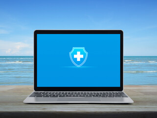 Cross shape with shield flat icon on modern laptop computer screen on wooden table over tropical sea and blue sky with white clouds, Business healthy and medical care insurance online concept