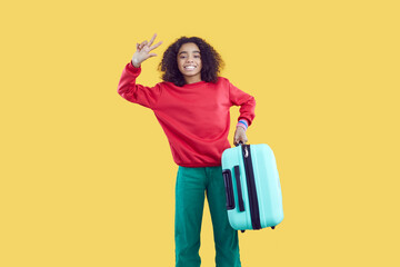 Children's tourism. Happy cute african american teenage girl holding travel suitcase isolated on yellow background. Joyful curly ethical kid girl tourist in casual clothes posing showing V-sign.