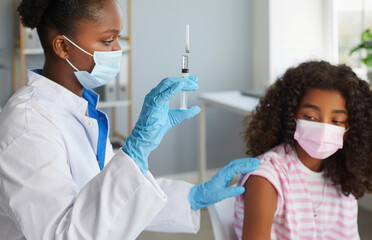 Doctor african american woman is giving a vaccine injection to teen girl in shoulder in clinic. Healthcare, vaccination from flu, covid-19, immunization concept. They wearing protective medical masks.