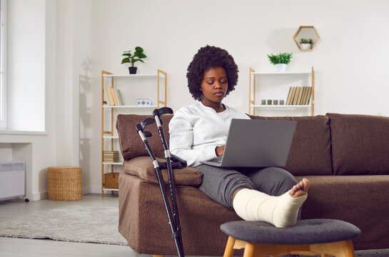Attractive African American woman with broken leg in cast is sitting on the sofa in the living room with laptop in hands. Recovery and rest at home after rehabilitation of broken leg. Crutches nearby.
