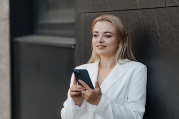 Blond stylish woman at oversized white jacket using phone on the modern city background
