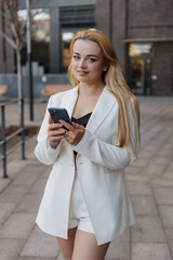 Obraz premium Blond stylish woman at oversized white jacket using phone on the modern city background