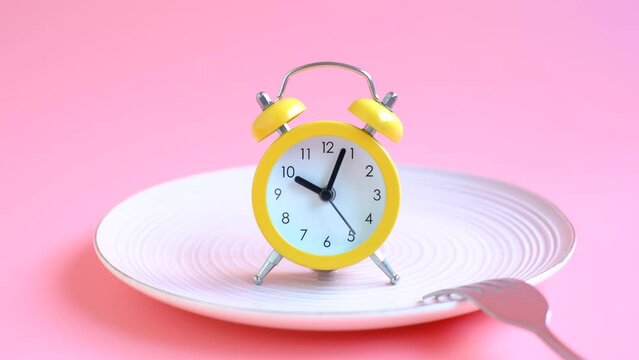 Empty plate with alarm clock and fork on pink background, intermittent fasting concept.
