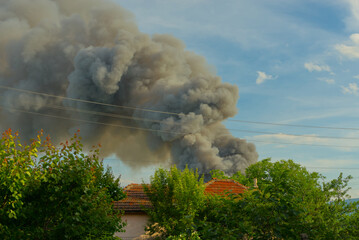 A huge fire with a tall black plume of smoke against a blue sky and green fields