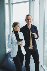 Smiling man and woman standing near window with gadgets in modern transparent office. Conversation of business colleagues in an informal setting.