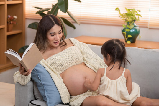 young pregnant mother reading book to her toddler daughter at home,.Image of attractive family pregnant woman and her little daughter smiling and reading book while sitting on sofa at home.