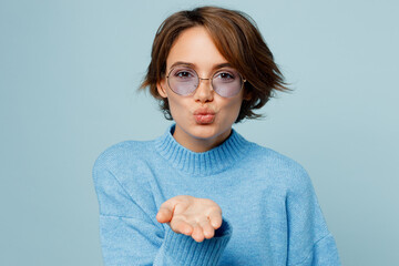 Young smiling happy cheerful fashionable fun caucasian woman wear knitted sweater glasses blow air kiss isolated on plain pastel light blue cyan background studio portrait. People lifestyle concept