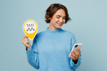Young smiling happy cheerful caucasian woman wear knitted sweater use mobile cell phone booking taxi cab isolated on plain pastel light blue cyan background studio portrait. People lifestyle concept.