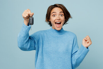 Young caucasian woman wear knitted sweater look camera hold in hand car key fob keyless system do winner gesture isolated on plain pastel light blue cyan background studio. People lifestyle concept.