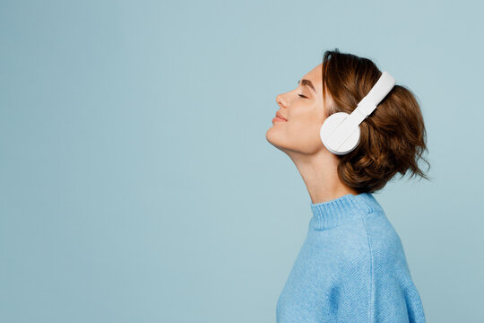 Young happy fun caucasian woman wear knitted sweater headphones listen to music with closed eyes have fun isolated on plain pastel light blue cyan background studio portrait. People lifestyle concept.