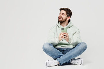 Full body smiling young man wear mint hoody look camera hold in hand use mobile cell phone look aside on workspace isolated on plain solid white background studio portrait. People lifestyle concept.