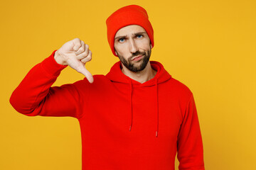 Fototapeta premium Young displeased dissatisfied sad caucasian man wear red hoody hat look camera showing thumb down dislike gesture isolated on plain yellow color background studio portrait. People lifestyle concept.