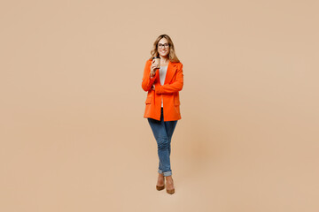 Full body young employee business woman corporate lawyer wear classic formal orange suit glasses work in office hold takeaway delivery craft brown cup coffee to go isolated on plain beige background.