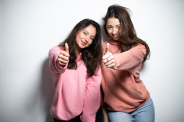 Two young attractive brunette girls in pink sweaters, smiling, showing like gesture with fingers, white background