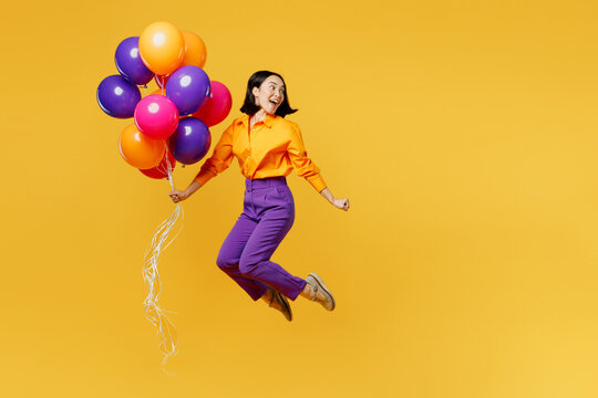 Full body side view happy fun young woman wear casual clothes celebrating hold bunch of balloons look aside on area jump high isolated on plain yellow background Birthday 8 14 holiday party concept