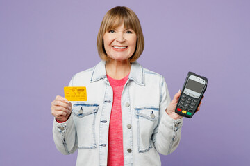 Elderly blonde woman 50s years old wear casual clothes denim jacket t-shirt hold wireless modern bank payment terminal to process acquire credit card isolated on plain pastel light purple background.