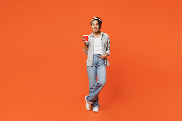 Fototapeta na wymiar Full body smiling fun young woman of African American ethnicity she wears grey shirt headband hold takeaway delivery craft paper cup coffee to go isolated on plain orange background studio portrait.