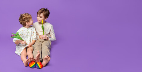 Fototapeta premium Children are sitting over the purple background with copy space for design advertisement. Full body view of smiling children in casual beige clothes. Colored rainbow hearts and tulips.