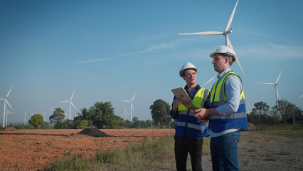 Two engineers men working at wind turbine farm. They discuss use tablet stand near wind turbines ecological energy industry power windmill field worker renewable energy. Clean energy concept.