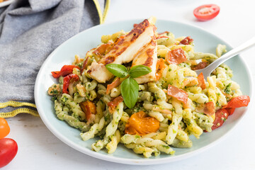 Fresh fusilli pasta with green pesto, baked tomatoes, crunchy ham and grilled cheese served on white plate