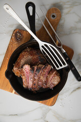 Entrecote Steak vom Rind medium rare gebraten in Gusseisen Pfanne mit Meersalz auf Holzbrett mit Messer, Fleischgabel und hellen Marmor Hintergrund