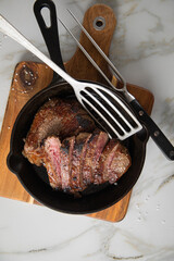 Entrecote Steak vom Rind medium rare gebraten in Gusseisen Pfanne mit Meersalz auf Holzbrett mit Messer, Fleischgabel und hellen Marmor Hintergrund