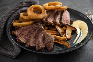 Entrecote Steak vom Rind medium rare gebraten mit Zwiebelringe, Pommes und Sauce Hollandaise auf schwarzen Steingut Teller mit Gabel, auf dunklen Stein Hintergrund