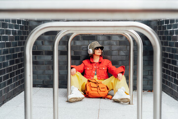 Hipster fashion young woman in bright clothes, sun glasses, bucket hat and earphones sitting near...