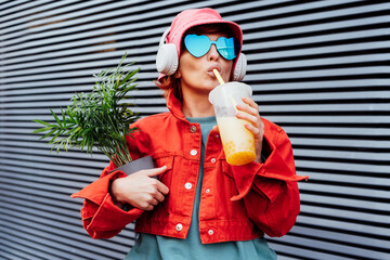 Hipster fashion woman in bright clothes, heart shaped glasses, headphones, bucket hat drinking...