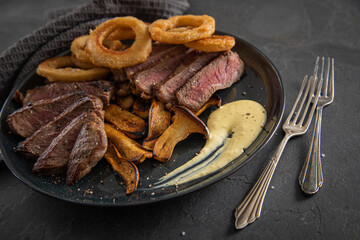 Entrecote Steak vom Rind medium rare gebraten mit Zwiebelringe, Pommes und Sauce Hollandaise auf schwarzen Steingut Teller mit Gabel, auf dunklen Stein Hintergrund