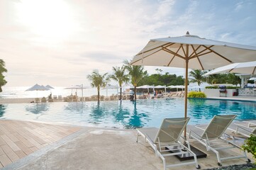 White sand beach and blue sea landscape view of a tropical resort hotel beside modern swimming pool, white parasols, palm trees, deck beach chairs in the golden sunset time