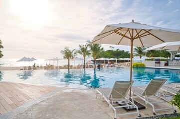 White sand beach and blue sea landscape view of a tropical resort hotel beside modern swimming pool, white parasols, palm trees, deck beach chairs in the golden sunset time