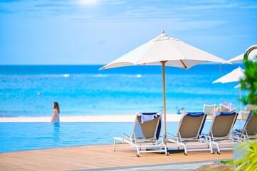 White sand beach and blue sea landscape view of a tropical resort hotel beside modern swimming pool, white parasols, palm trees, deck beach chairs in the golden sunset time