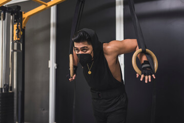 Fototapeta na wymiar A masked asian man leaning on wooden still rings. A gymnast preparing to train at the gym.