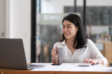 Charming Asian businesswoman sitting in the office with a digital laptop computer. Excited Asian businesswoman raising hands to congratulate while working in a modern office,