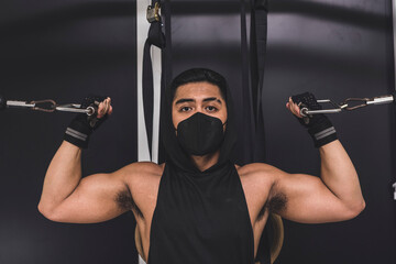 Fototapeta na wymiar A masked asian man does standing overhead bicep cable curls. Wearing a black low cut tank top. Training arms at the gym.