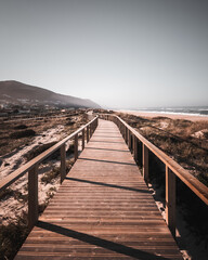 Fototapeta na wymiar A long boardwalk on a grassy sand dune at the beach leading to the mountain and ocean with desaturated colors. The way ahead concept, Quiaios Beach, Portugal