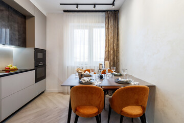 Modern kitchen combined with the dining room in dark and light colors, with orange chairs and glass cabinets for dishes