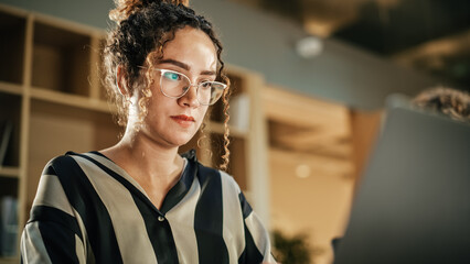 Portrait of Hispanic Inspired Young Woman Smiling and Using Laptop in Bright Contemporary Office. Female Client Advisor Answering Customer Support E-mails, Eyes Focused on the Screen. Close Up Shot.