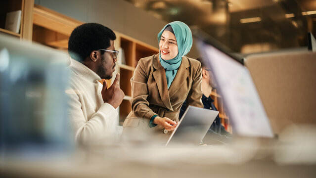 Two Coworkers Collaborating And Celebrating Their Accomplishment In A Modern Office. Muslim Female Trainer Using Laptop To Onboard A Black Male Customer Support Agent And Answering His Question.