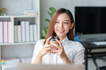 Pretty Asian businesswoman sitting happily working on her laptop and drinking coffee smiling and laughing brightly in the office.