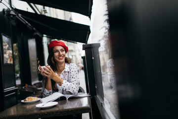 Young stylish woman in red beret having a french breakfast with coffee and croissant sitting...