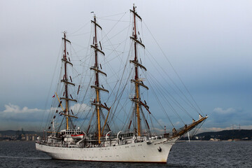 Fototapeta na wymiar Three-masted sailing frigate goes to sea from the port against a stormy sky.