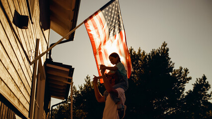 Happy Patriotic Dad Holding His Young Daughter of His Shoulders, Helping Her to Put the United States of America Flag on the Wall of Their House to Celebrate a National Holiday. Sunny Day in USA.