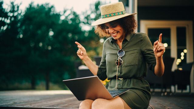 Portrait of a Positive Multiethnic Female Using Laptop Computer on a Porch of a House. Young Female, Sitting, Dancing, Browsing Internet and Ordering Stuff Online for a Relaxing Garden Party.