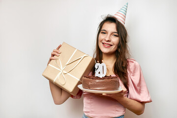 The girl is holding a festive cake with a candle in the form of the number 20 and a gift. Birthday celebration concept.